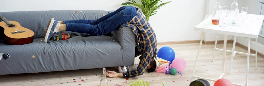 Mann ist verkatert und hängt von Couch runter, um ihn Luftballone und ein weißer Tisch auf hellem Hintergrund