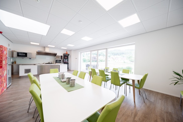 Salle lumineuse avec chaises vertes, grandes tables, kitchenette et vue sur l'extérieur par des fenêtres.