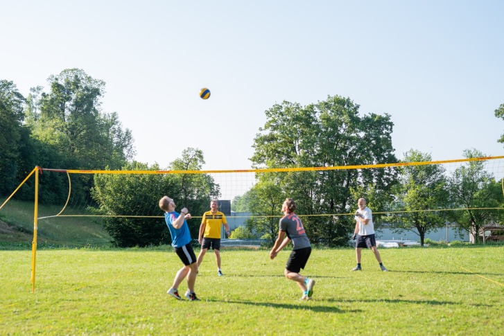 Quatre hommes jouent au volley-ball sur un terrain en herbe avec un filet, entouré d'arbres.