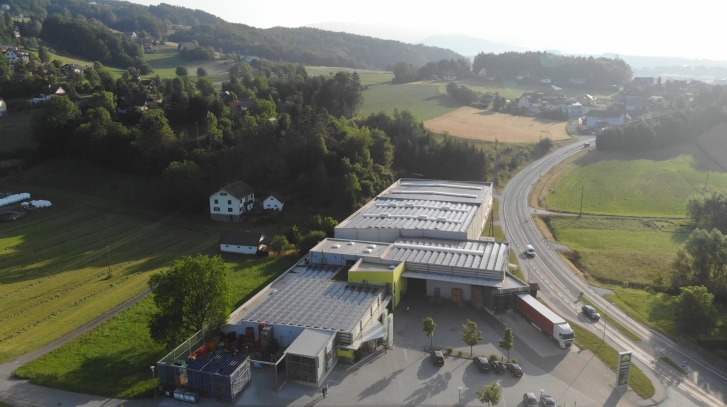 Bâtiment avec toits solaires, entouré de champs et d'arbres, situé sur une route avec des voitures.