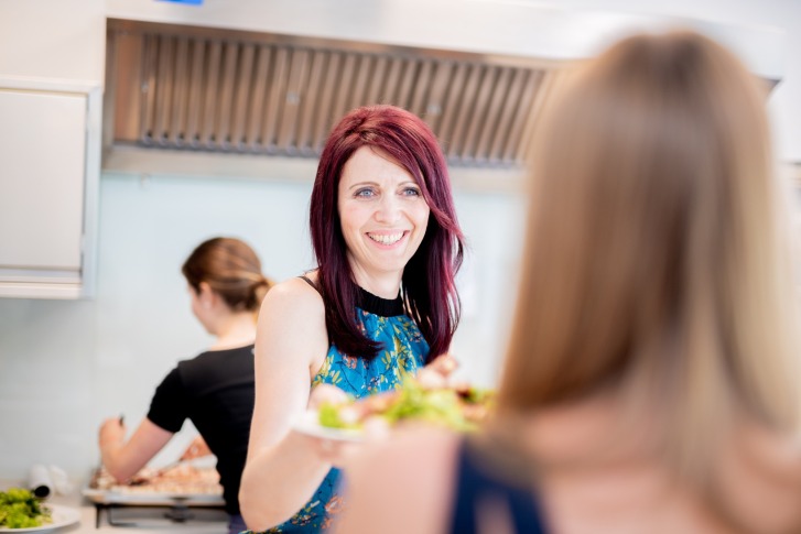 Une femme passe des assiettes à une autre personne en souriant, dans une cuisine moderne et lumineuse.