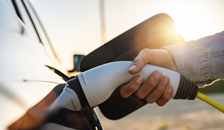 Hand holding charging cable plugged into car outside at sunset.