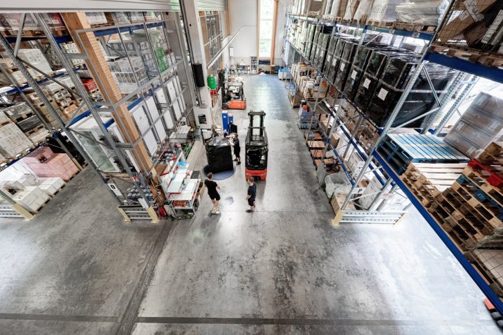 Shelves with beverage cartons are in a warehouse, forklift trucks move black pallets.