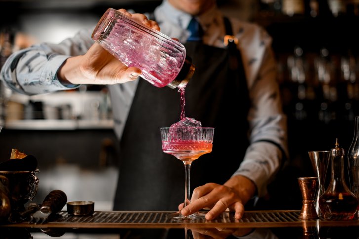 Een persoon schenkt een roze cocktail uit een shaker in een glas, staand in een baromgeving.