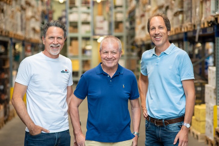Three men smile in a warehouse with shelves full of boxes in the background.