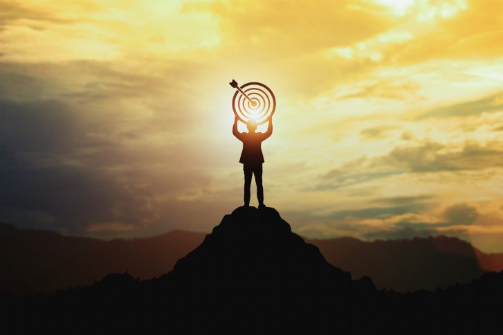Person holding target with arrow. Standing on a mountain peak in front of a colorful sunset sky.