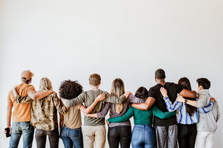 A group of people stand embraced in a row in front of a blank white wall.