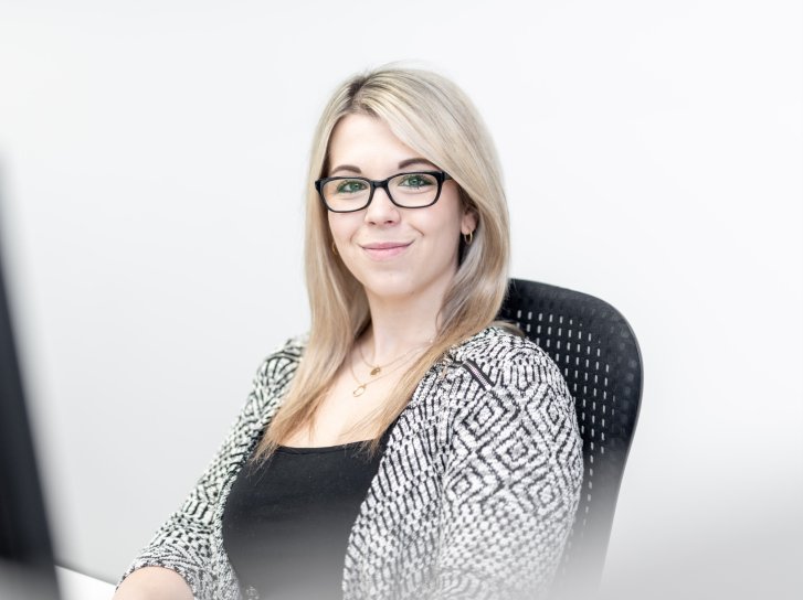 Eine blonde Frau mit Brille sitzt lächelnd in einem Bürostuhl vor hellem Hintergrund.