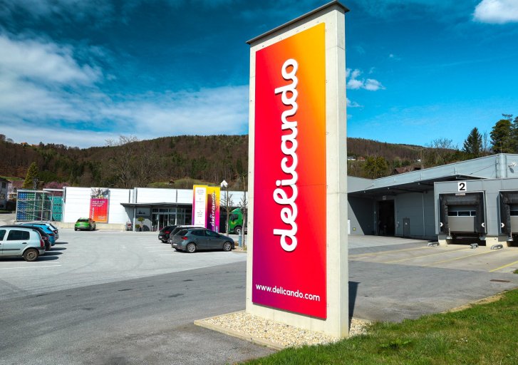 Groot, kleurrijk bord met het "delicando" logo staat voor een modern gebouw in een landelijke omgeving.
