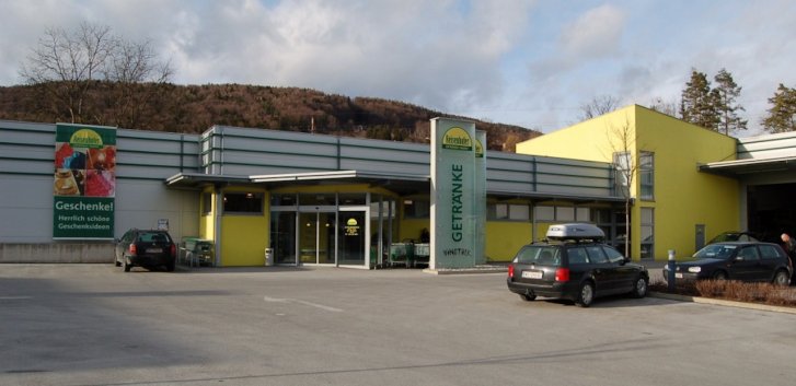 Gebäude steht ruhig auf Parkplatz, umgeben von Autos und Bäumen, mit Aufschrift "GETRÄNKE".