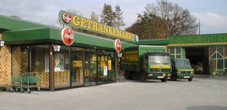 Magasin de boissons, des camions verts sont garés devant le bâtiment. Publicité pour Coca-Cola. Arbres en arrière-plan.