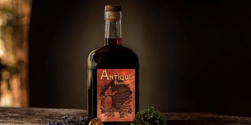 Bottle with "Antique Pelinkovac" inscription stands on a wooden background, surrounded by herbs and dried fruit.