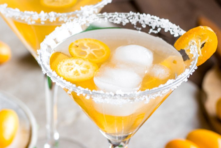 Cocktailglas mit Eis und Kumquats steht auf Tisch, umgeben von geschnittenen Früchten und Messer.
