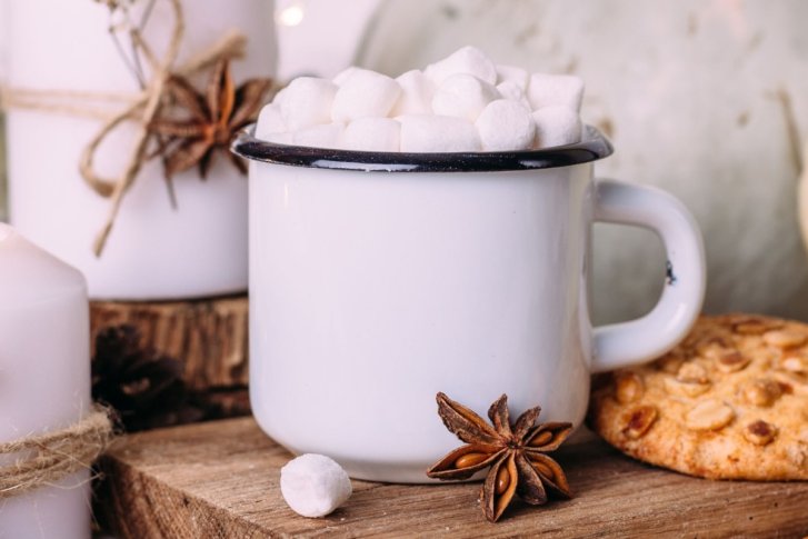 Weiße Tasse enthält Marshmallows, steht neben Keksen auf Holzbrett mit Kerzen und Dekorationen.