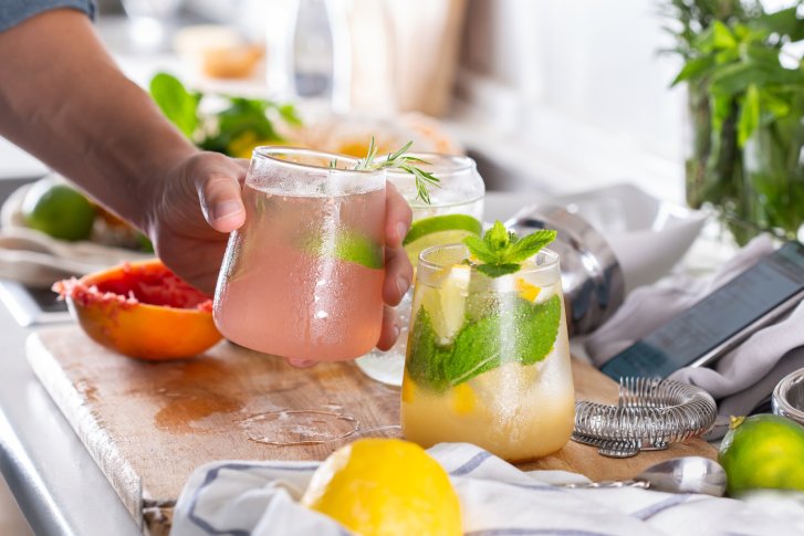Gläser mit Cocktails, auf einem Holzbrett, umgeben von Obst und Barzubehör.