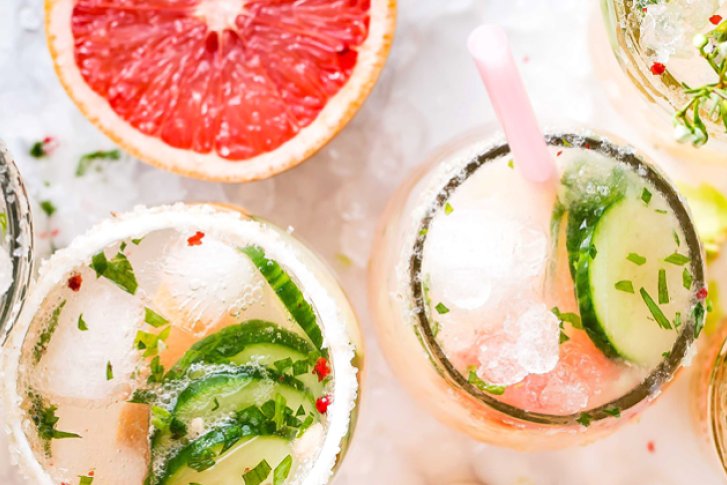 Gläser mit Cocktails und Gurken, auf Eis, dekoriert mit Kräutern und Grapefruitscheiben auf Marmoroberfläche.