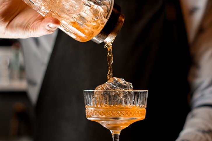 Hand gießt Getränk in elegantes Glas, Hintergrund zeigt verschwommene Barumgebung.