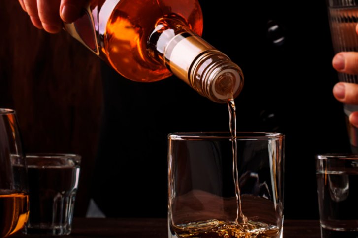 Eine Hand gießt goldene Flüssigkeit aus einer Flasche in ein Glas, umgeben von anderen Gläsern.