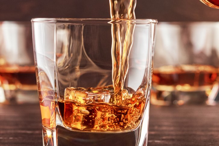 Whiskey wird in ein Glas mit Eiswürfeln gegossen, weitere Gläser im Hintergrund.