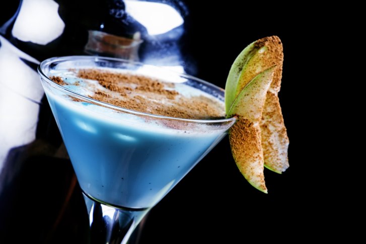 Blaues Getränk in Cocktailglas, dekoriert mit Apfelscheiben, im schwarzen Hintergrund.