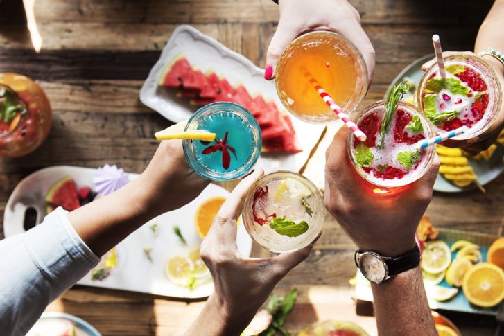 Bunte Cocktails werden von Händen über einem Holztisch mit tropischen Dekorationen und Früchten gehalten.
