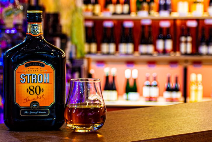 Flasche "Stroh 80" steht auf Theke neben Glas, im Hintergrund Regale mit Flaschen.