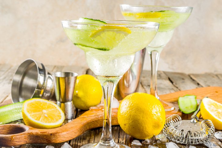 Zwei Cocktails stehen auf einem Holztisch, umgeben von Zitronen, Gurken und Barzubehör.