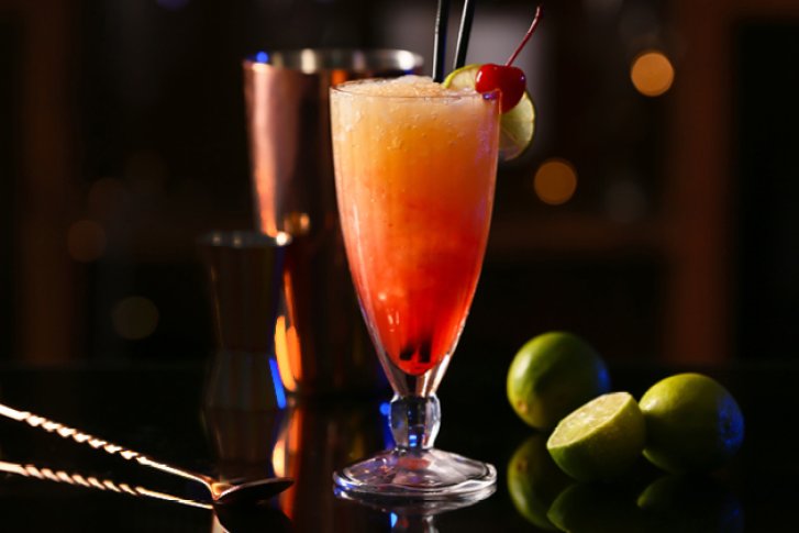 Cocktailglas mit Früchten dekoriert, steht auf schwarzem Tisch; Limetten und Barzubehör im Hintergrund.