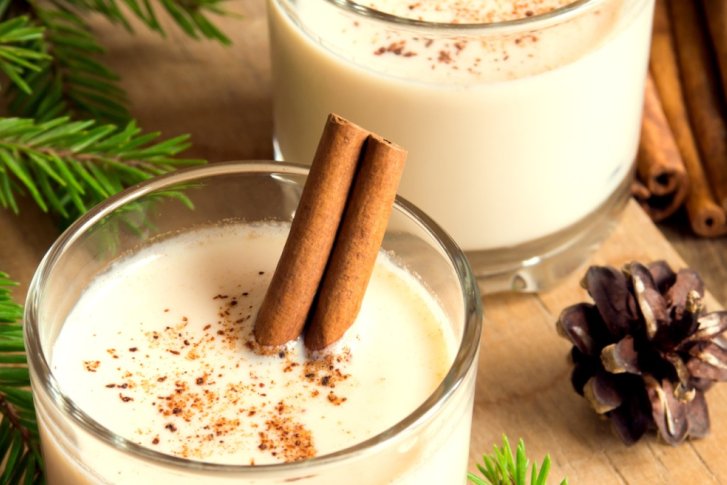 Gläser mit Eierlikör, dekoriert mit Zimtstangen, auf Holztisch mit Tannenzweigen und Zapfen.