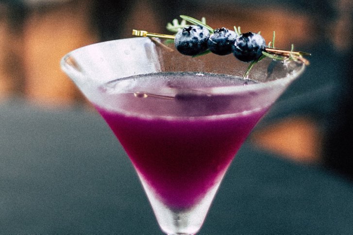 Lila Cocktail mit Blaubeeren-Garnitur in einem Martini-Glas, vor unscharfem Hintergrund.