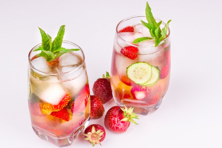 Zwei Gläser mit Erdbeeren, Gurkenscheiben und Minze in klarer Flüssigkeit auf weißem Hintergrund.