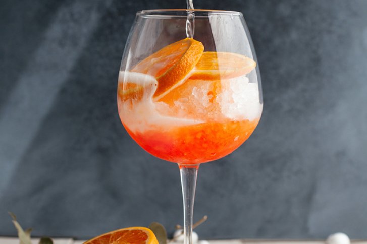 Ein Glas Orangentrunk mit Eis wird gefüllt, umgeben von geschnittenen Orangen, grauer Hintergrund.