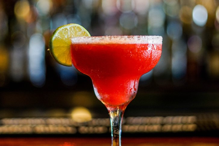 Roter Cocktail mit Limettenscheibe am Glasrand in einer Bar mit verschwommenem Hintergrund.
