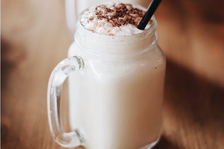 Ein gläserner Krug enthält einen cremigen Drink mit Kakaopulver und Strohhalm auf Holztisch.