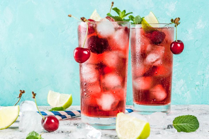 Zwei hohe Gläser mit rotem Getränk, Eis und Kirschen stehen auf einem Tisch mit Limetten.