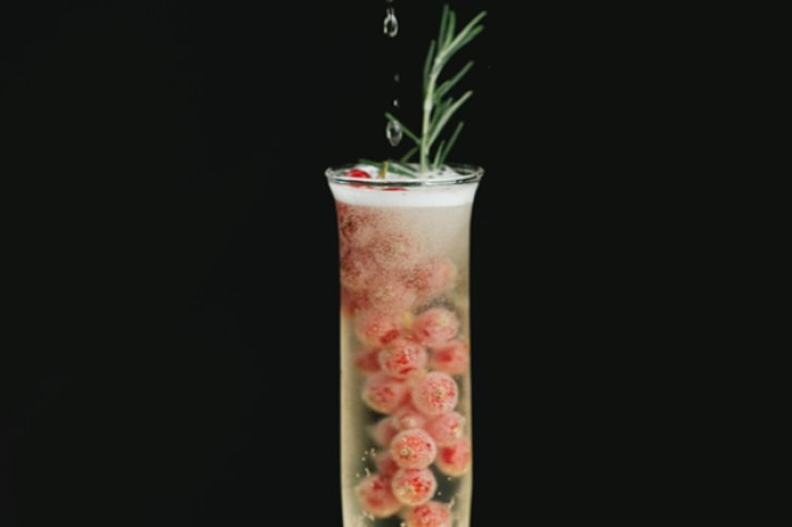 Ein Sektglas mit Rosmarin und roten Beeren, daneben tropft Flüssigkeit, vor schwarzem Hintergrund.