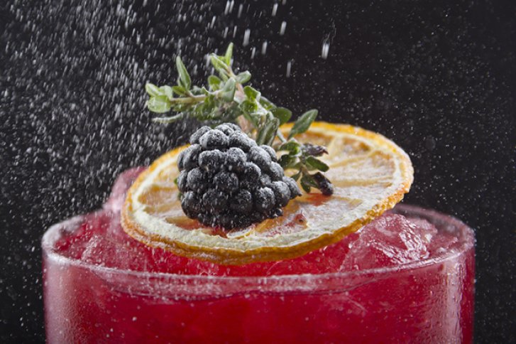 Ein rotes Getränk wird mit Brombeere und Limette garniert, umgeben von herabfallendem Zucker.