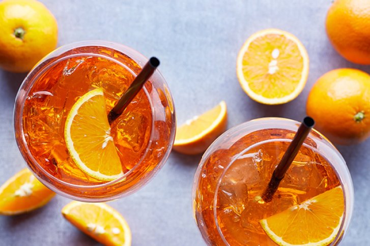 Zwei Cocktails mit Orangenscheiben und Strohhalmen, umgeben von frischen Orangen auf grauem Hintergrund.