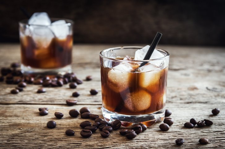 Zwei Gläser mit Eiskaffee und Strohhalmen stehen auf Holztisch, umgeben von Kaffeebohnen.