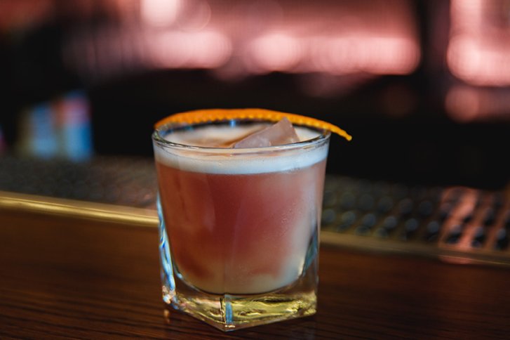 Ein Cocktailglas mit Eiswürfeln und Orangenscheibe steht auf einer hölzernen Baroberfläche.