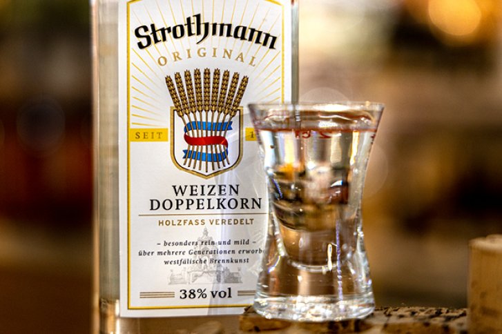 Ein Glas klarer Schnaps steht vor einer Strothmann Weizen Doppelkorn Flasche im Hintergrund.