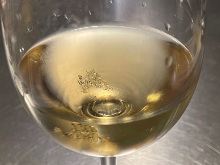Ein Glas mit weißem Getränk, innen schwimmen kleine Kristalle, auf metallischer Oberfläche.