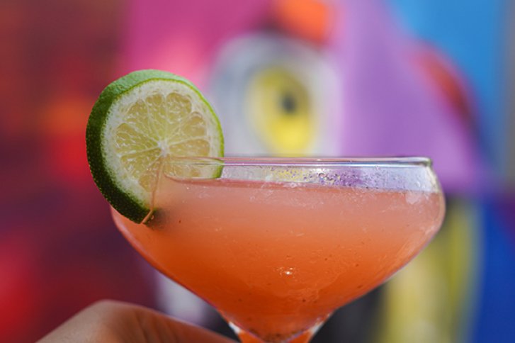 Ein rosa Cocktail mit Limettenscheibe dekoriert, vor einem bunten, unscharfen Hintergrund gehalten.