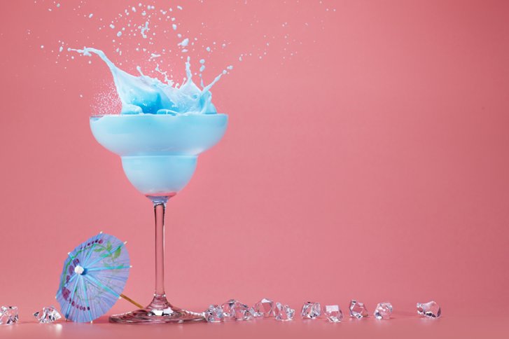 Blaues Getränk spritzt im Cocktailglas. Rosa Hintergrund, Eiswürfel und Papier-Regenschirm daneben.