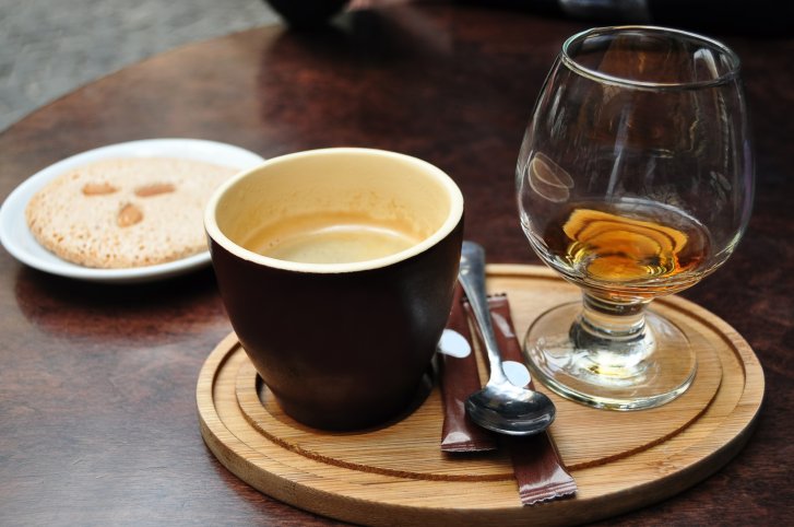 Espressotasse steht neben Glas mit Cognac auf Holztablett, vor Teller mit Keks.