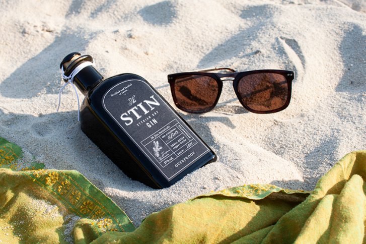 Schwarze Gin-Flasche liegt im Sand neben einer Sonnenbrille, umgeben von grünem Tuch.