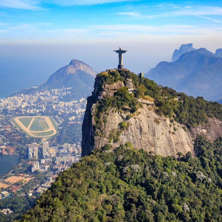 Statue stands on mountain peak, with city, mountains and sea in the background.
