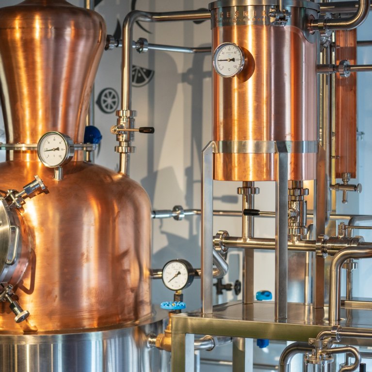 Koperen destillatietoestel in een brouwerij, omringd door pijpen en meetinstrumenten.