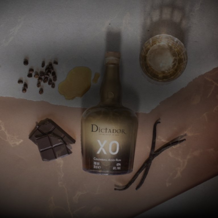 Een productfoto van bovenaf, op ooghoogte, toont een fles XO likeur omringd door chocolade, koffiebonen, vanillestokjes en een glas op een bruine stenen ondergrond.