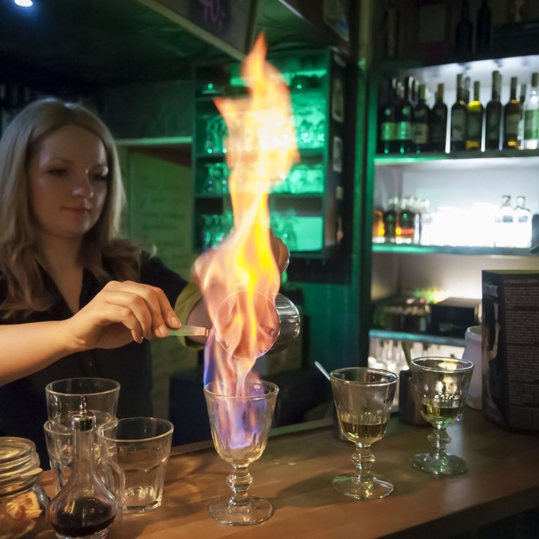 Woman flambéing drink behind bar counter while glasses and bottles are illuminated in the background.